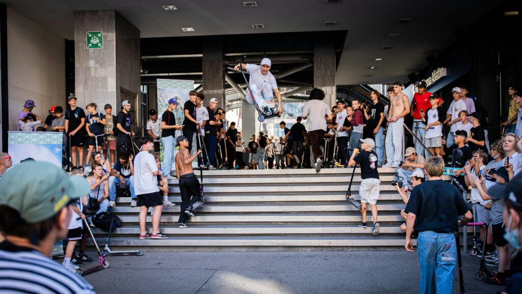 Exciting street scene featuring a scooter stunt jump with an engaged crowd, showcasing youth culture and extreme sports.